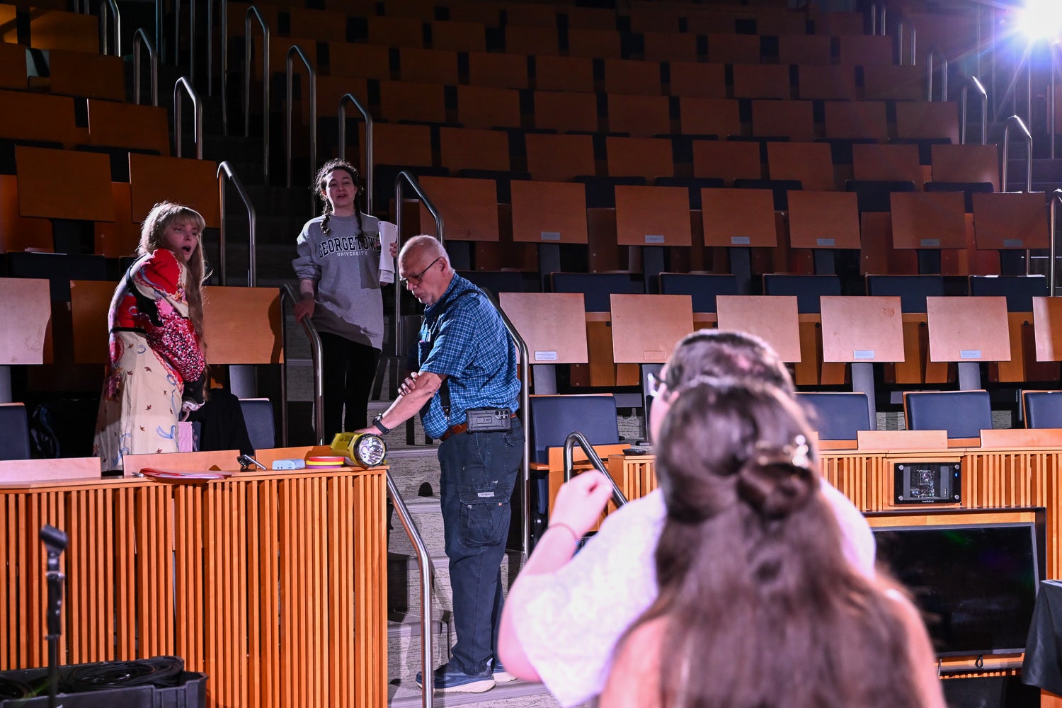 People standing in tiered auditorium seating speak, as actors on the stage in the foreground put on their costumes