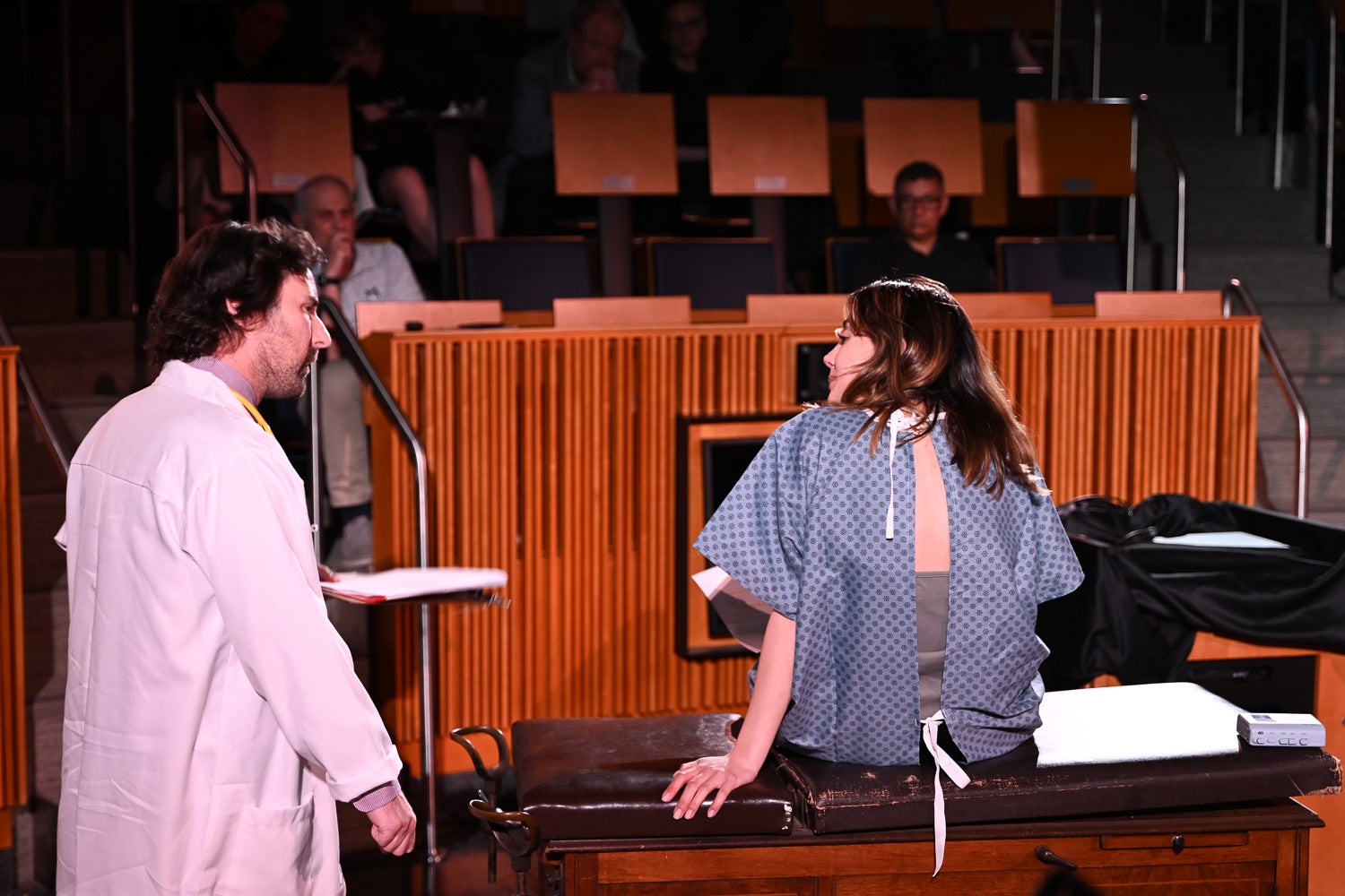 Sofi Fateh, wearing a hospital gown, sits on the exam table listening to John E. Loughlin-Presnal while acting in April Brassard's