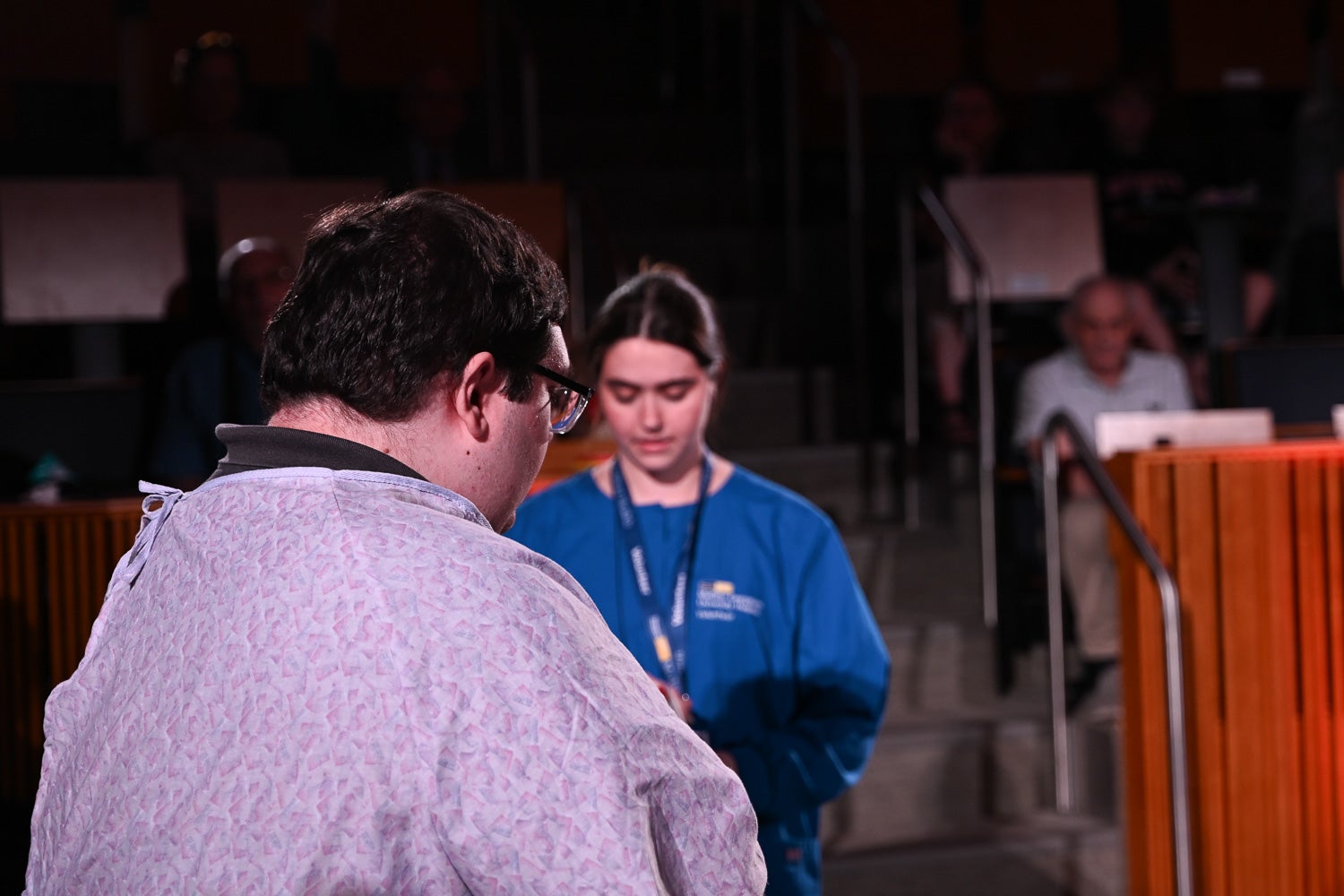 Alexandros Georgussis, wearing a hospital robe, sits on the exam table on the Harvey Amphitheater stage in front of Catherine Hughes, wearing a MedStar shirt, while acting in