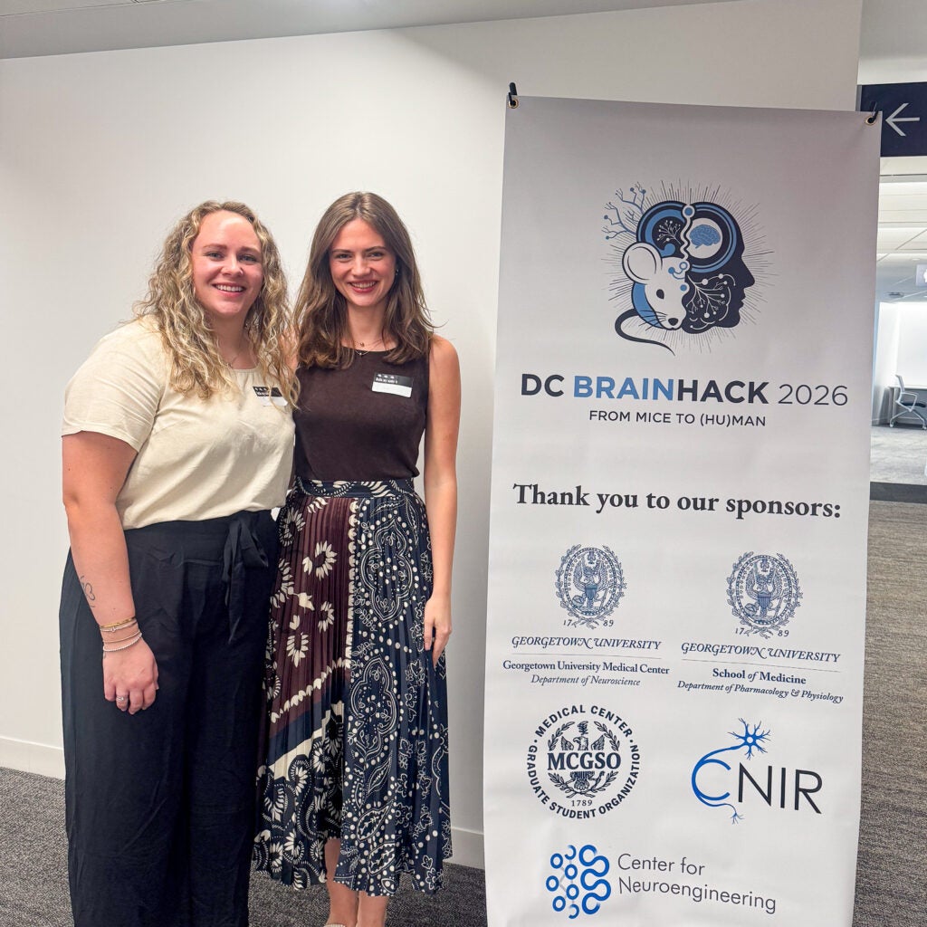 Peyton Subia and Chloe Casagrande pose next to a sign for "DC Brainhack 2026" which lists event sponsors