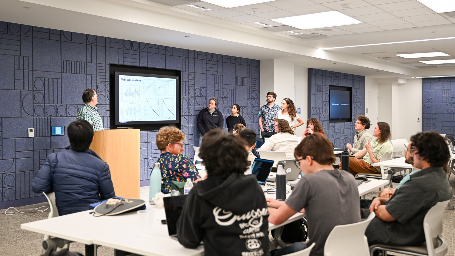 A team presents a slide on a big screen, while audience members sat at tables listen