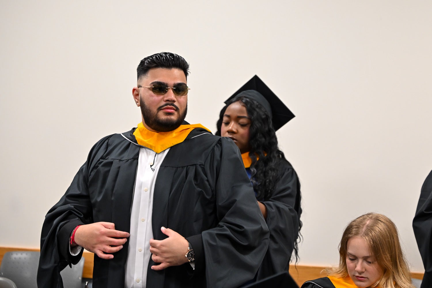 A graduate wearing sunglasses gets help with his regalia hood from a peer in the line-up room