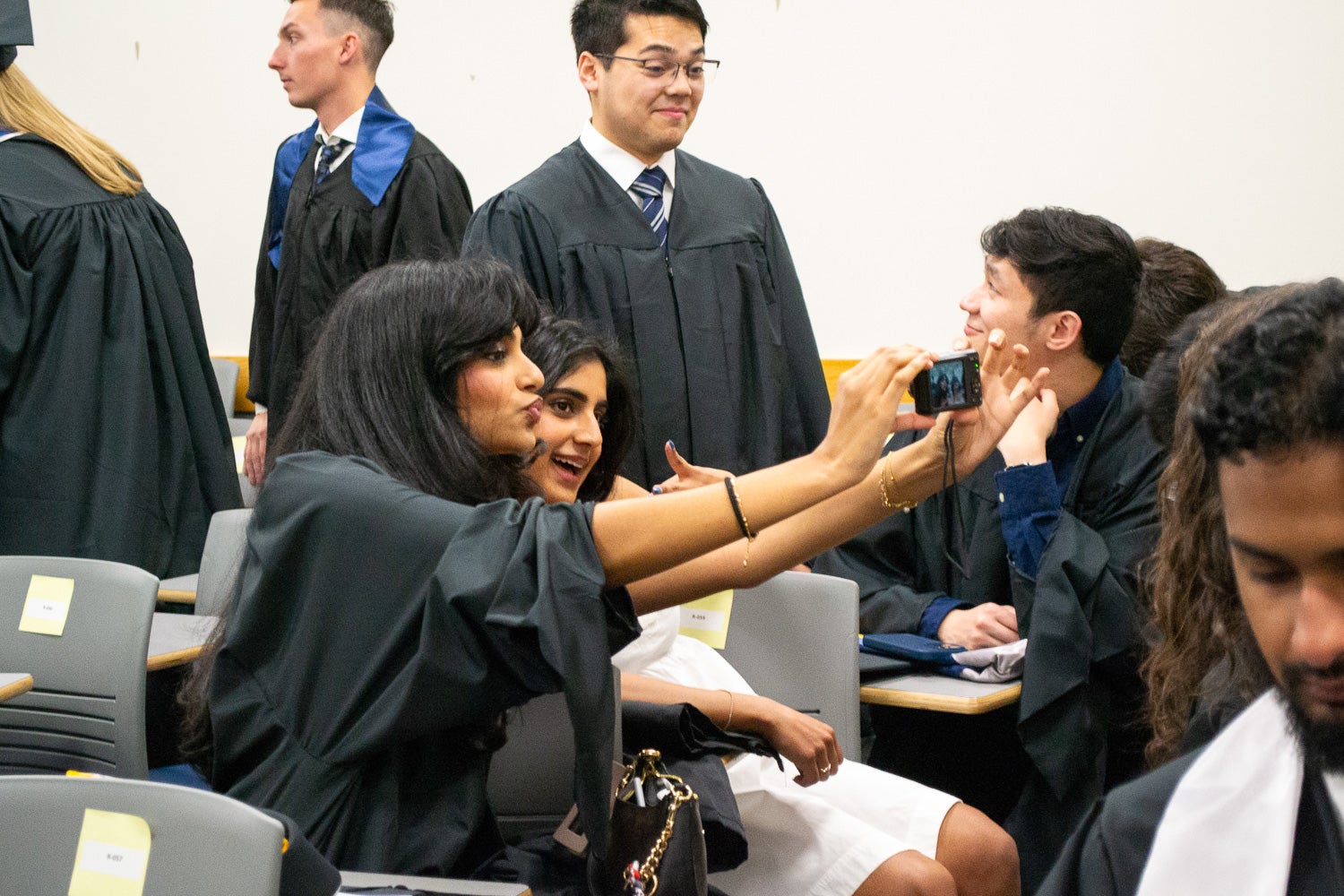 Two graduates dressed in commencement regalia take a selfie together in the line-up room