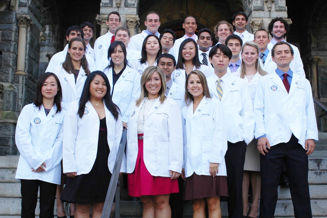 Students wearing white coats from the Georgetown University School of Medicine pose together on the steps of Healy Hall