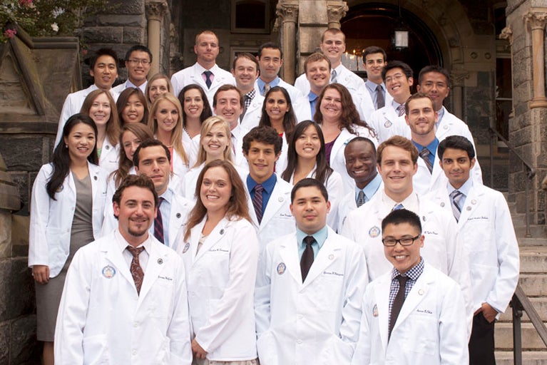Students wearing white coats from the Georgetown University School of Medicine pose together on the steps of Healy Hall