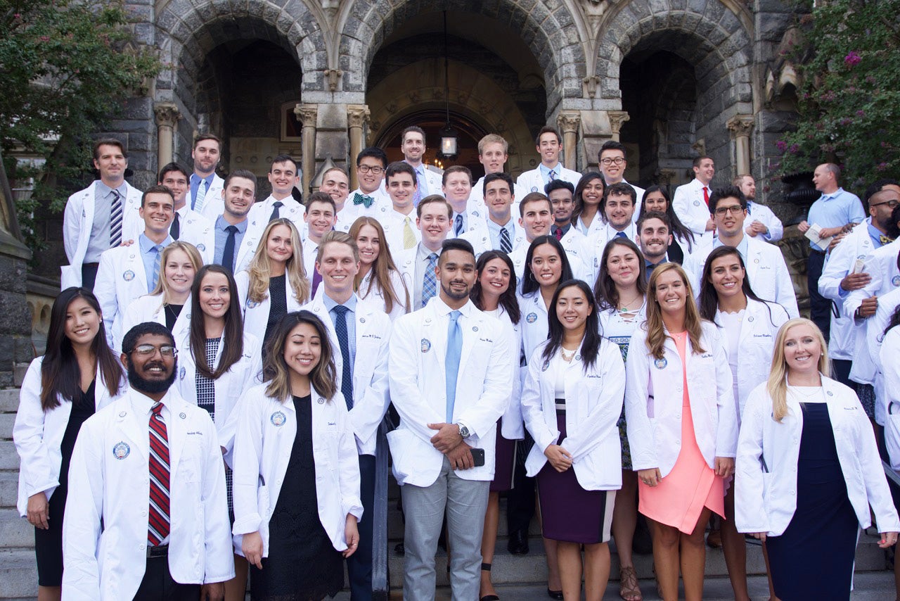 Students wearing white coats from the Georgetown University School of Medicine pose together on the steps of Healy Hall
