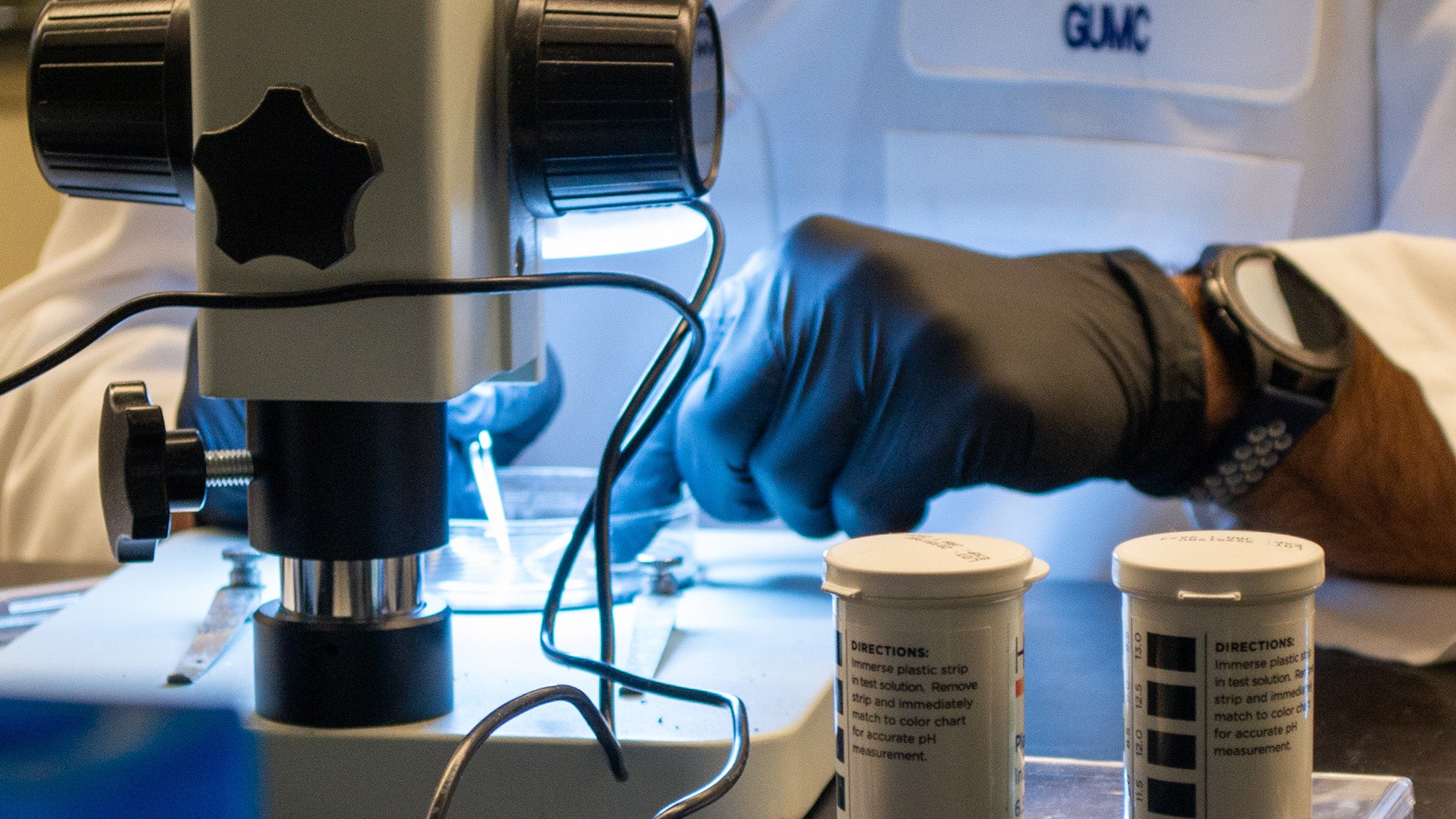 A doctoral student wearing a lab coat and gloves uses a microscope